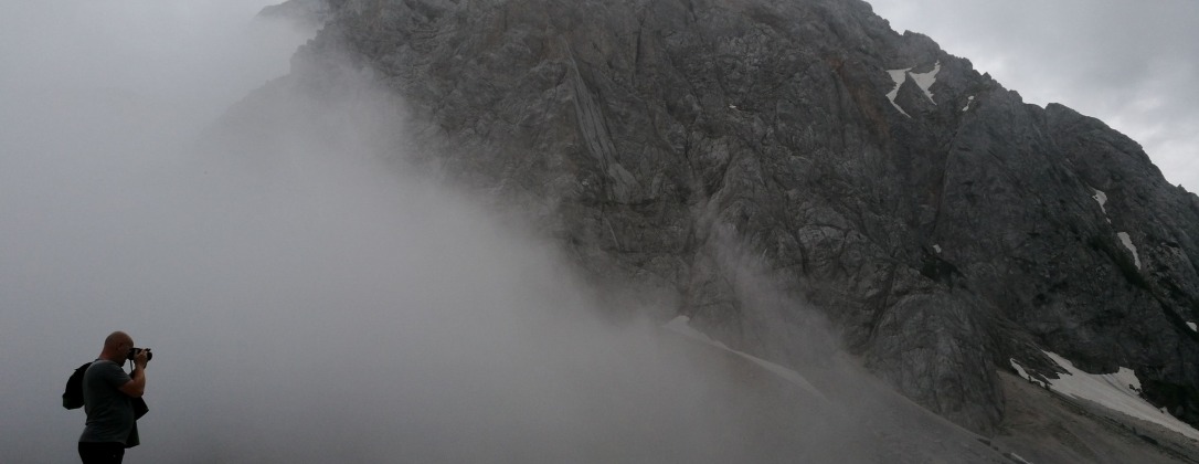 a man staring into a foggy mountain from its smaller peak.
